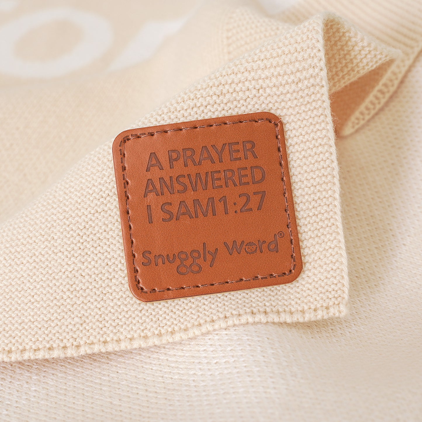 Close-up of cream knit baby blanket with brown leather tag, "A PRAYER ANSWERED I SAM 1:27" and "Snuggly Word".