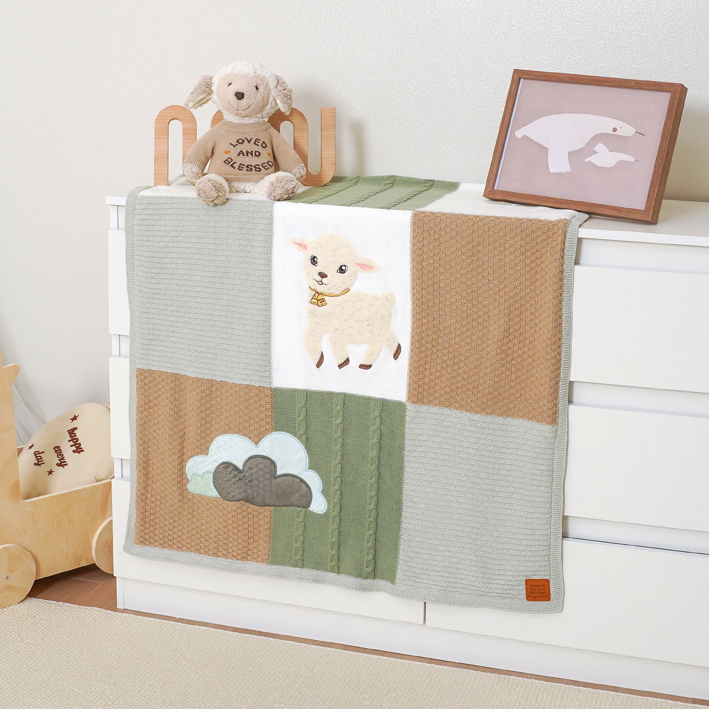 Goodness & Mercy Psalm 23 knitted baby blanket with lamb & clouds, draped on a white dresser in a nursery.
