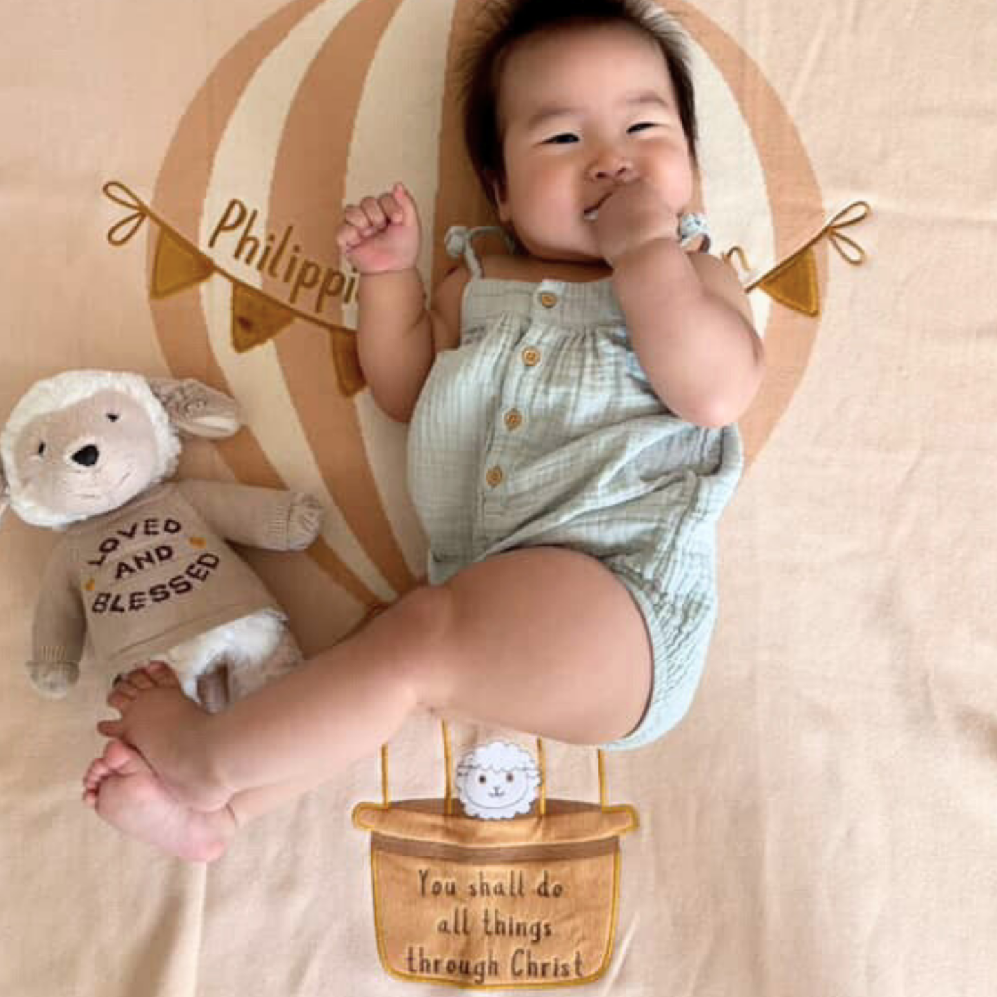 Smiling baby on "Soar High Philippians 4:13" milestone blanket with hot air balloon, beside "Loved and Blessed" lamb.